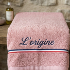 Serviette de bain rose pastel en coton éponge épais avec la broderie personnalisée 'L’origine' en bleu marine cursive, ornée d'une bande tricolore bleu, blanc, rouge, posée sur une table en bois avec un flacon d'huile en arrière-plan.