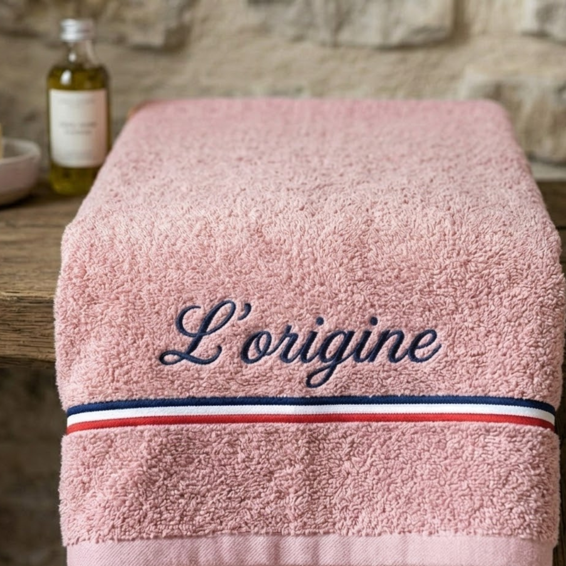 Serviette de bain rose pastel en coton éponge épais avec la broderie personnalisée 'L’origine' en bleu marine cursive, ornée d'une bande tricolore bleu, blanc, rouge, posée sur une table en bois avec un flacon d'huile en arrière-plan.