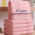 Serviette de bain rose pastel en coton éponge épais avec la broderie personnalisée 'L’origine' en bleu marine cursive, ornée d'une bande tricolore bleu, blanc, rouge, posée sur une table en bois avec un flacon d'huile en arrière-plan.