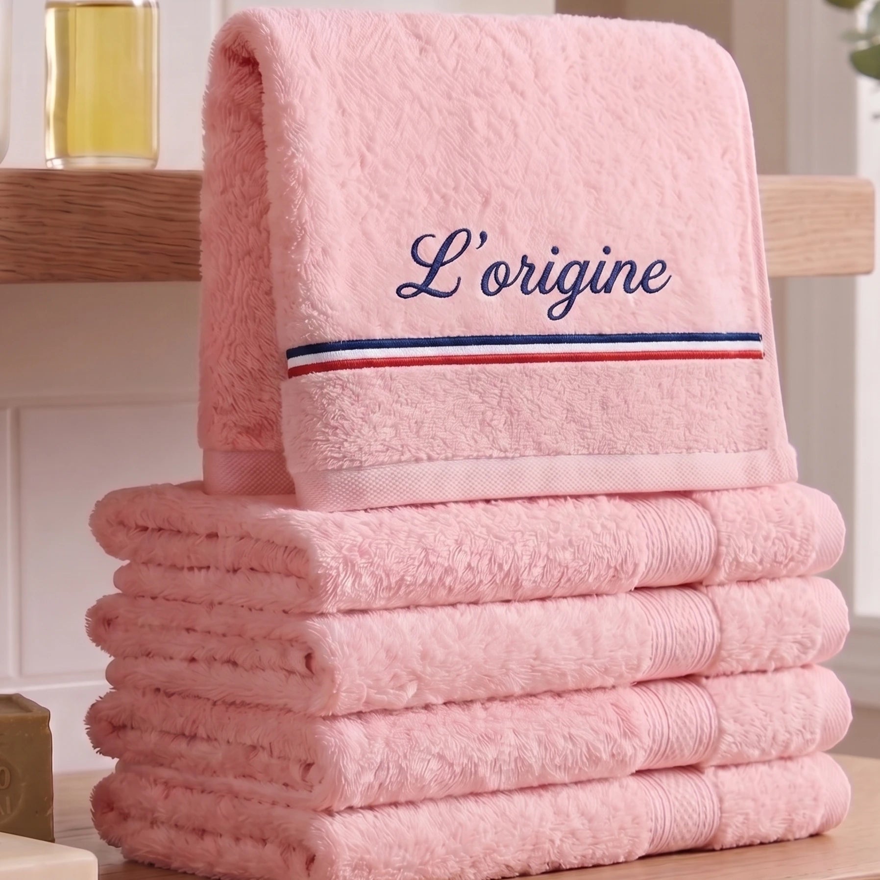 Serviette de bain rose pastel en coton éponge épais avec la broderie personnalisée 'L’origine' en bleu marine cursive, ornée d'une bande tricolore bleu, blanc, rouge, posée sur une table en bois avec un flacon d'huile en arrière-plan.