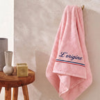 Serviette de bain rose pastel en coton éponge épais avec la broderie personnalisée 'L’origine' en bleu marine cursive, ornée d'une bande tricolore bleu, blanc, rouge.