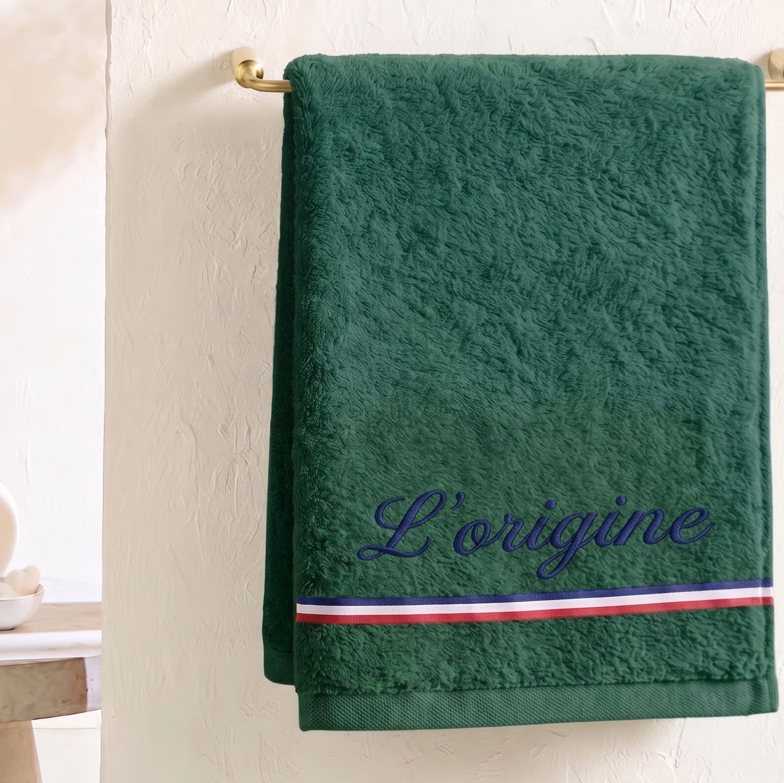 Serviette de bain verte en coton éponge avec une broderie bleu marine cursive 'L’origine' et une bande décorative tricolore (bleu, blanc, rouge), suspendue sur un porte-serviette.