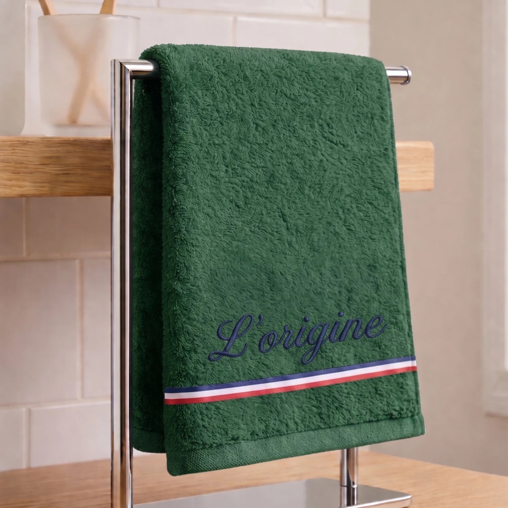 Serviette de bain verte en coton éponge avec une broderie bleu marine cursive 'L’origine' et une bande décorative tricolore (bleu, blanc, rouge), suspendue sur un porte-serviette.