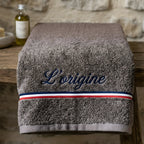 "Une serviette de bain haut de gamme de couleur marron, portant la broderie élégante 'L’origine' en bleu. Le design inclut une bande tricolore française (bleu, blanc, rouge) distinctive. La serviette est présentée pliée sur un meuble en bois rustique dans un décor chaleureux de salle de bain."