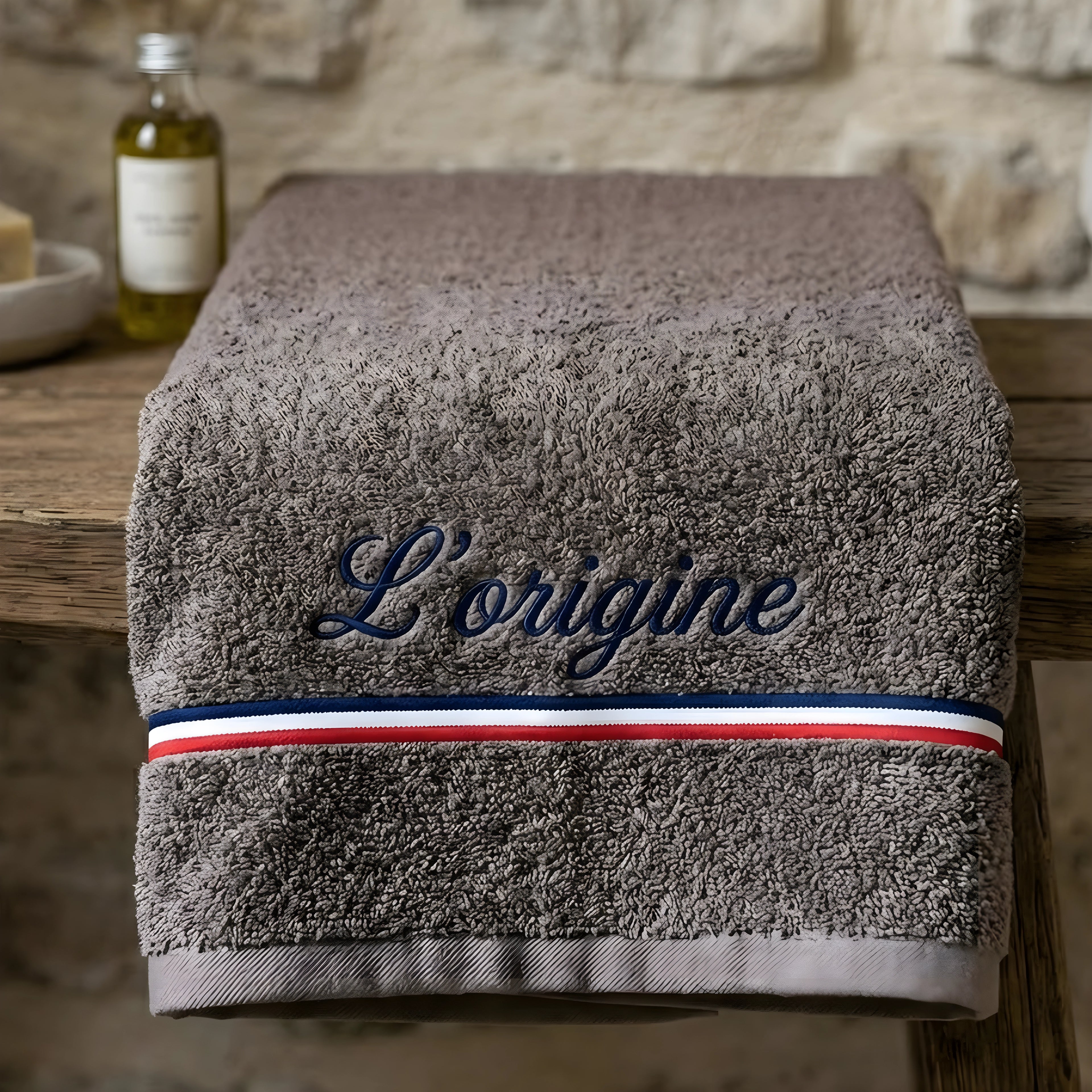 "Une serviette de bain haut de gamme de couleur marron, portant la broderie élégante 'L’origine' en bleu. Le design inclut une bande tricolore française (bleu, blanc, rouge) distinctive. La serviette est présentée pliée sur un meuble en bois rustique dans un décor chaleureux de salle de bain."