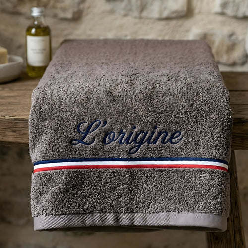 "Une serviette de bain haut de gamme de couleur marron, portant la broderie élégante 'L’origine' en bleu. Le design inclut une bande tricolore française (bleu, blanc, rouge) distinctive. La serviette est présentée pliée sur un meuble en bois rustique dans un décor chaleureux de salle de bain."